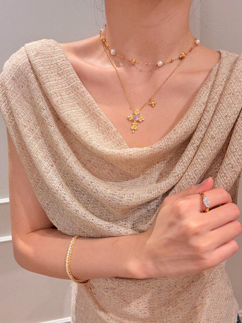 Person wearing a gold necklace with a cross pendant, gold bracelet, and ring on a neutral background
